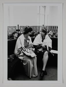 Two women sitting on a bench, wearing prayer shawls and kippot and holding prayer books