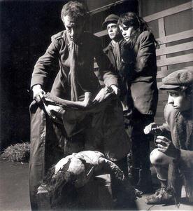 Four actors in tattered clothing. One man drapes a coat over a dummy while the others watch
