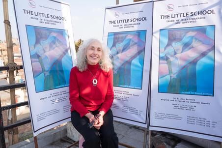 photograph of a woman with white curly hair wearing a read sweater in front of posters for her play "The Little School"