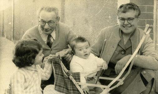 black and white photograph of a little girl with dark hair and a baby in a stroller with an older man and woman