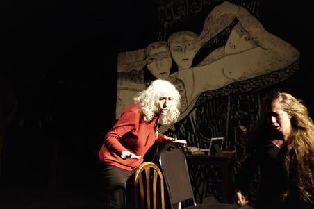photograph of a woman with white curly hair wearing a red sweater and a woman in black with long brown hair acting, in front of a set with abstract haunted faces