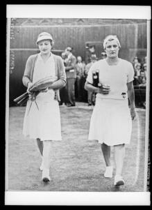 Black and white photograph of two women tennis players in white skirts carrrying tennis rackets, walking towards the camera