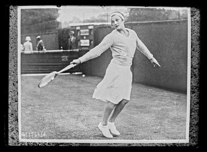 black and white photograph of a woman in a white skirt playing tennis, with outstretched arms