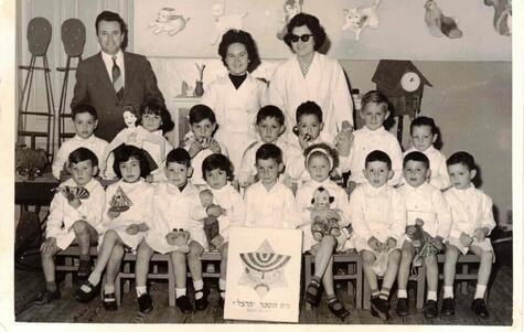 black and white photograph of a daycare with two seated rows of small children dressed in white with three adults standing