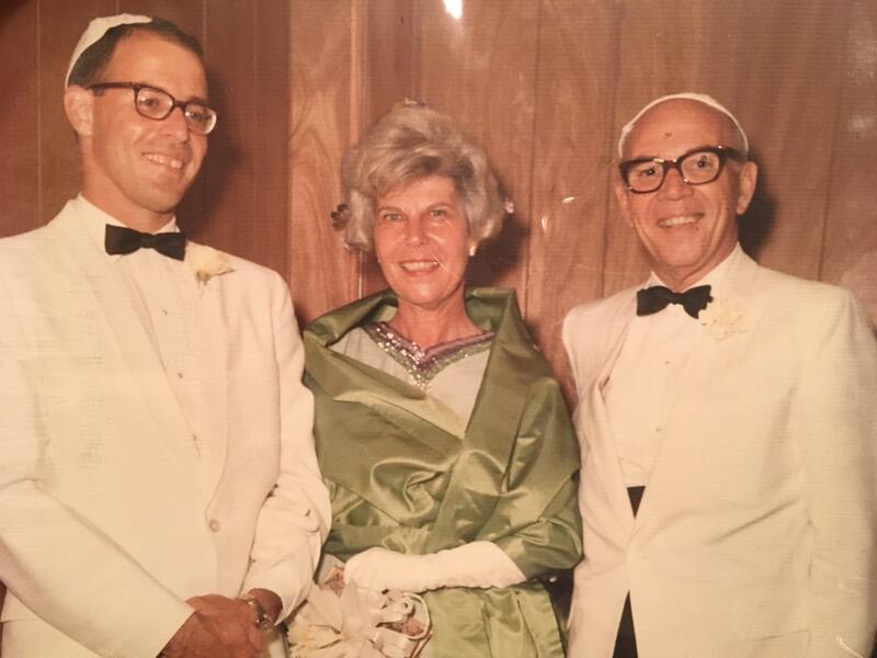 A smiling trio in formal attire stands against a wood-paneled background. Two men wear white suits and kippahs; the woman in the center dons a green dress.