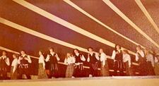 A group of dancers performing onstage, with the women wearing long dresses and headscarfs, and the men wearing black vests, hats, and trousers, white shirts, and tallit katan