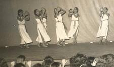 A group of six women, barefoot, wearing long white tunics, and holding up clay pots as they dance onstage