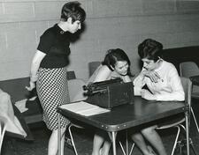 Black and white photo of three women at a table with a typewriter