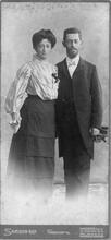 historical black and white photograph of a woman in a white blouse and long dark skirt and a man wearing a dark suit with a long jacket