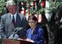 Ruth Bader Ginsburg speaking at a podium outdoors, with Bill Clinton standing behind her and an American flag in the background