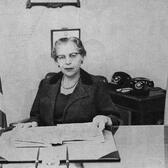 Anna Moscowitz Kross sitting at a desk with a file open in front of her. In the background are two telephones, a diploma on the wall, and the flags of the United States and New York City
