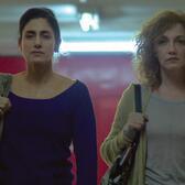 A woman with dark hair in a bun in a blue top, left, and a woman with curly blonde hair stand side in fluorescent lit space. Both have right arm holding purses in place on shoulder. 