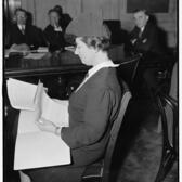 Dorothy Straus sitting a table, leafing through pieces of paper, with several men sitting in the background watching her