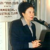 Ora Namir standing in front of a microphone with a sign behind her with Hebrew and English