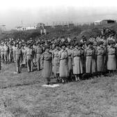 A large group of soldiers standing at attention in a rural area. At the front stand two lines of women soldiers in uniform dress and berets.