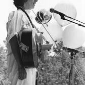 Black and white photograph of a woman with short hair at a microphone with a guitar, outdoors