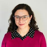 Betsy has long, curly, dark hair and light skin. She is wearing thin framed glasses, and a maroon sweater over a collared polkadot shirt. She is looking at the camera with a slight smile and is in front of a plain gray background.