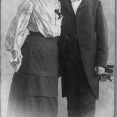 historical black and white photograph of a woman in a white blouse and long dark skirt and a man wearing a dark suit with a long jacket