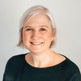 Jenny has medium length white hair and light skin. She is wearing a dark green top. She is smiling at the camera and is in front of plain light gray background. 