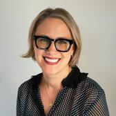 Mikki has medium length blond hair and light skin. She is wearing thick black glasses and a black polkadot shirt in front of a plain gray background. She is looking at the camera and smiling.
