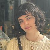 Sophie Haward stands in front of a shop window. She has curly dark hair cut in a bob with bangs, and is wearing a white cabled sweater. She is smiling at the camera.