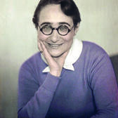 photograph of a seated woman wearing a purple sweater and round glasses, with her chin in her hand, smiling at the camera