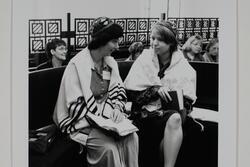 Two women sitting on a bench, wearing prayer shawls and kippot and holding prayer books