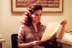 Ruth Behar sitting in a chair beside a desk, reading a piece of paper, beneath a large framed tapestry hanging on the wall