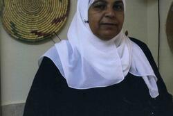Amna Kenanalk holding a program, with a woven decoration hanging on the wall behind her