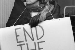 photograph of a woman with curly gray hair wearing a sign saying "End the Occupation"