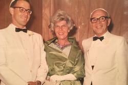 A smiling trio in formal attire stands against a wood-paneled background. Two men wear white suits and kippahs; the woman in the center dons a green dress.
