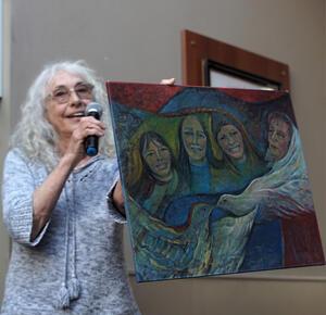 photograph of a woman with white curly hair in a blue sweater with a painting of four women