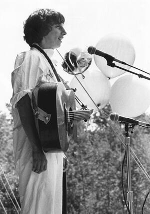 Black and white photograph of a woman with short hair at a microphone with a guitar, outdoors