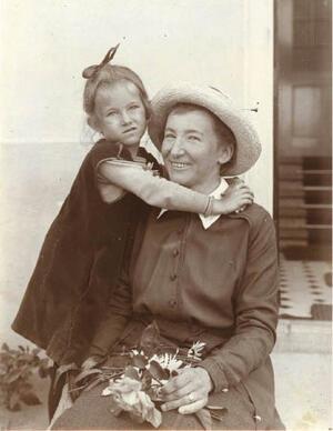 Black and white photograph of a smiling woman with a hat and holding flowers, with a young girl posed with her arms around the woman