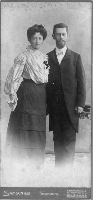 historical black and white photograph of a woman in a white blouse and long dark skirt and a man wearing a dark suit with a long jacket