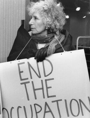 photograph of a woman with curly gray hair wearing a sign saying "End the Occupation"
