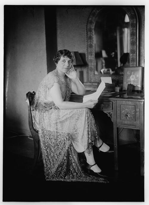 Estelle Liebgling, seated at a dressing table with a mirror and a portrait of a child, holding a folded piece of paper