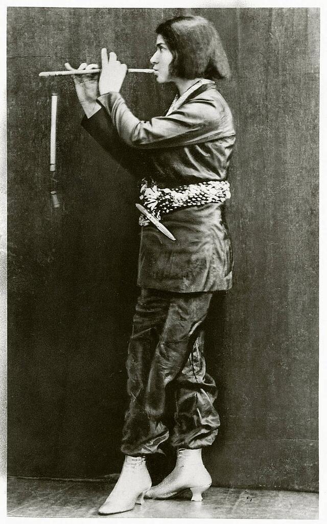 Else Lasker Schuler dressed in men's clothing with a decorative belt, playing a pipe