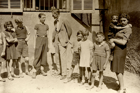 An adult man and woman standing in front of a house with a group of children