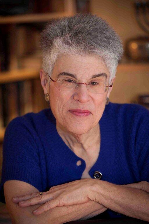 Judith Hauptman wearing a blue top and glasses smiling into the camera with arms crossed over one another