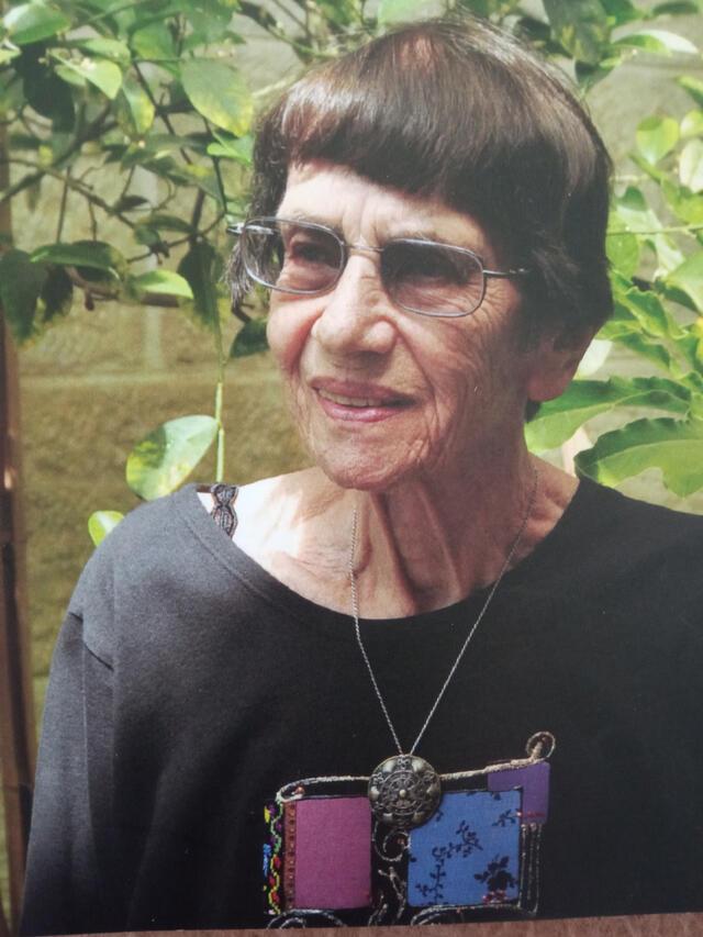 Color portrait of Judith Shuval against background of trees and plants and brick wall