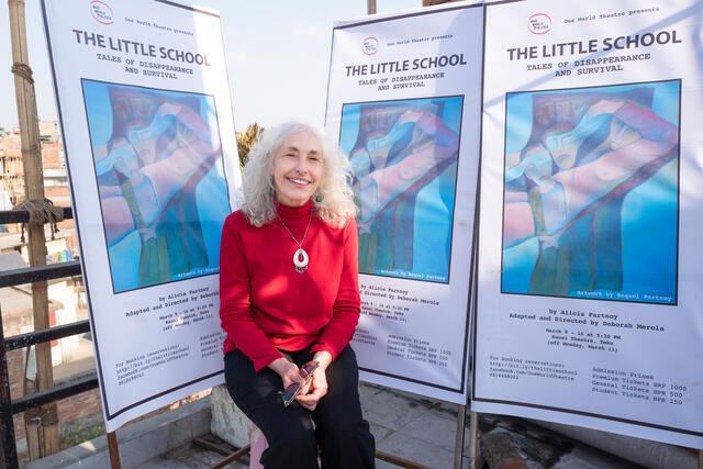 photograph of a woman with white curly hair wearing a read sweater in front of posters for her play "The Little School"