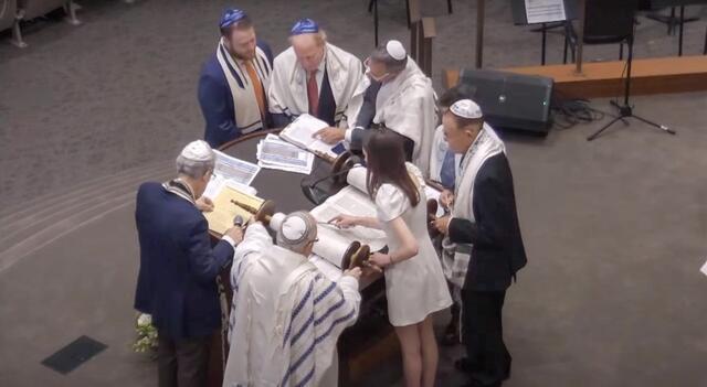 Annie Katz reading Torah at the Bimah surrounded by 6 men