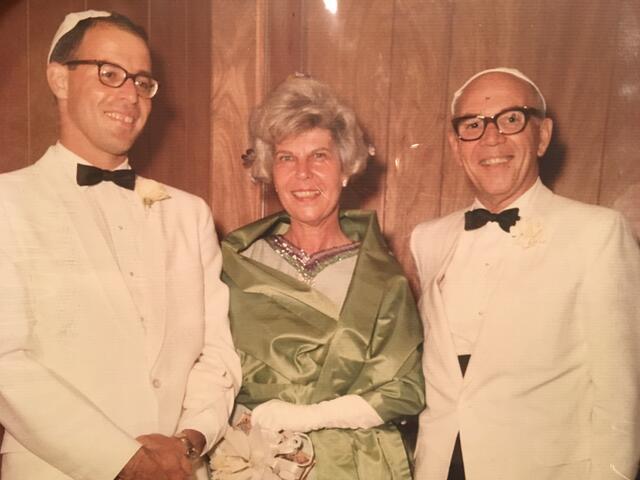 A smiling trio in formal attire stands against a wood-paneled background. Two men wear white suits and kippahs; the woman in the center dons a green dress.