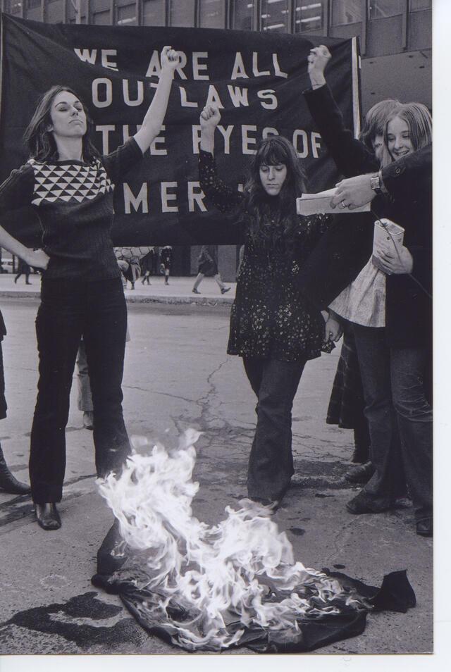 Black and white phogoraph of three women with their arms raised and defiant expressions, in front of burning judges robes