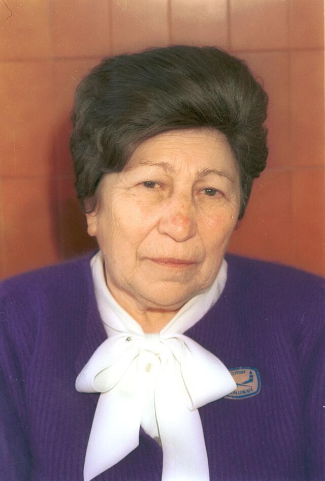 photograph of a woman with a dark wig wearing a blue suit with a white bow, looking unsmiling at the camera